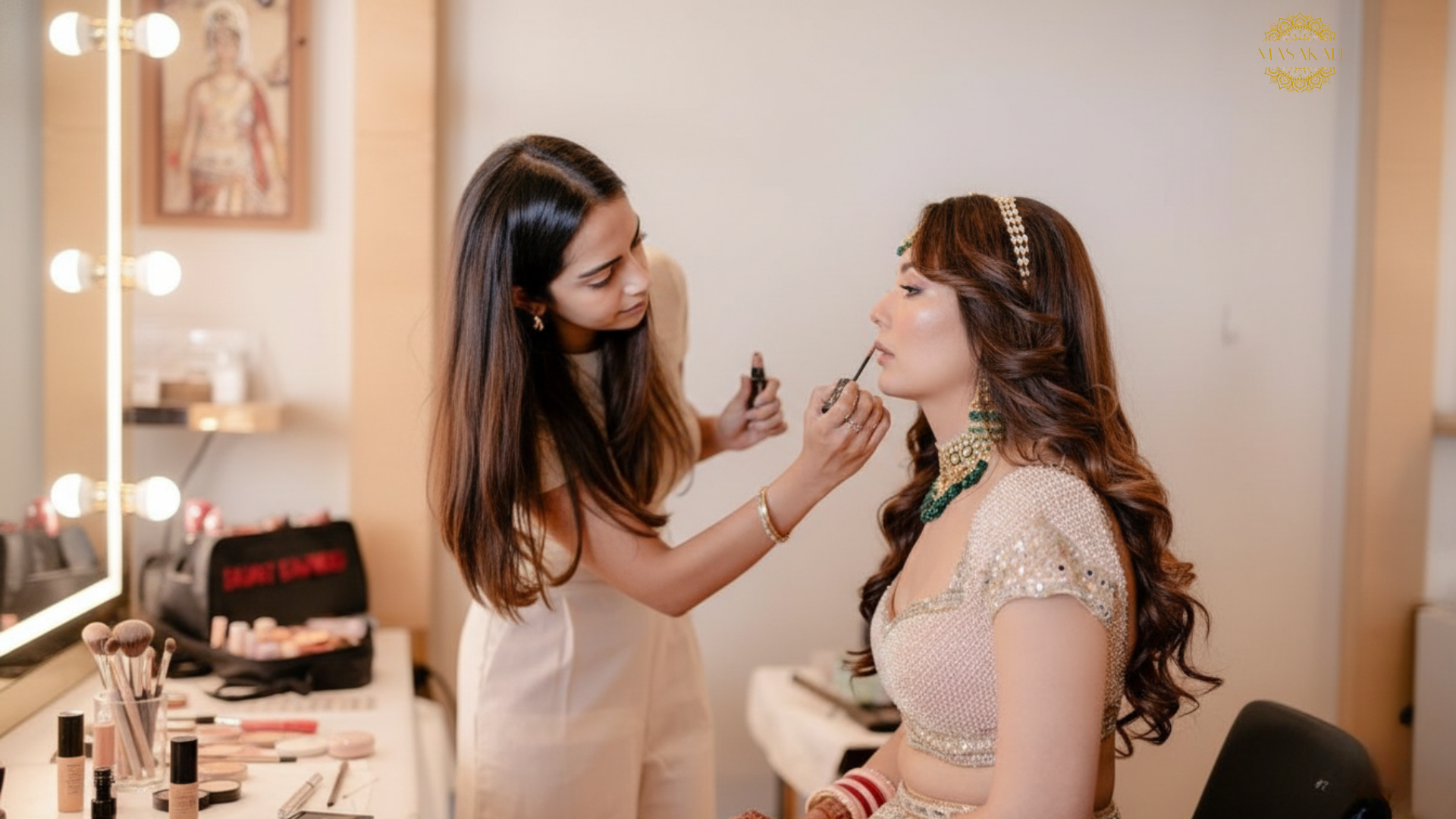 An Indian makeup artist applies lipstick to a bride, who is wearing a sparkling traditional lehenga and ornate jewelry, in a bright, modern studio with warm lighting and a vanity mirror.
